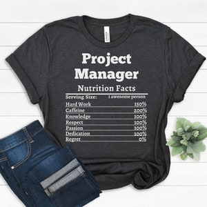 May include: Dark gray t-shirt with the words "Project Manager" and "Nutrition Facts" printed in white. The nutrition facts include percentages for hard work, caffeine, knowledge, respect, passion, dedication, and regret. Blue jeans are folded below the shirt.