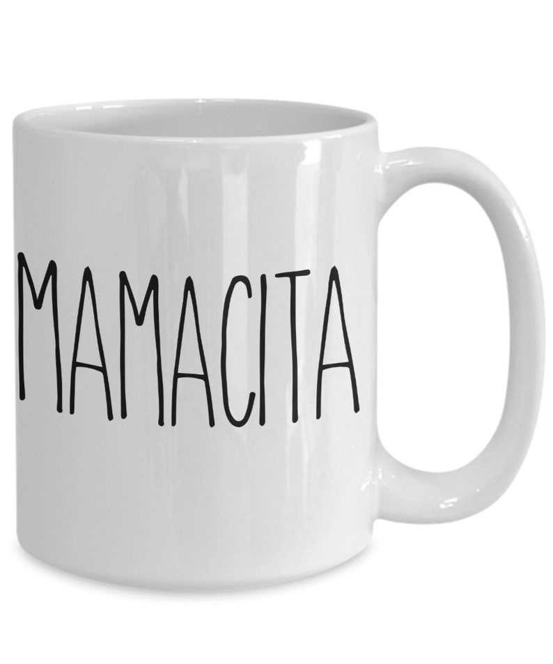 May include: White ceramic mug with the word "MAMACITA" printed in black, sans-serif font. The mug has a curved handle and a glossy finish. The mug is designed for hot or cold beverages.