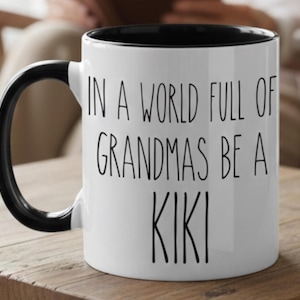 May include: A white ceramic mug with a black handle and black interior rim. The exterior features black text in a simple sans-serif font that reads: IN A WORLD FULL OF GRANDMAS BE A KIKI. The mug has a cylindrical shape and a smooth, glossy finish.