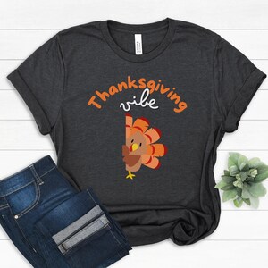 May include: Dark gray t-shirt with the words "Thanksgiving vibe" in orange and white. A cartoon turkey with orange and brown feathers peeks from the right side. The shirt is paired with blue jeans.
