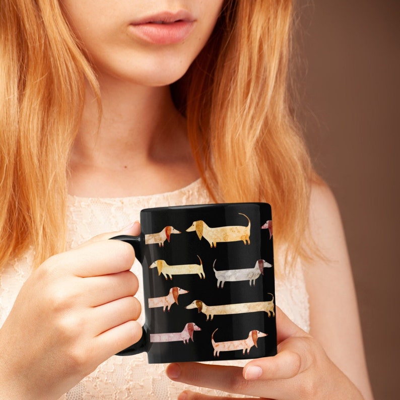 May include: Black ceramic mug featuring a repeating pattern of watercolor dachshund dogs in various colors. The dogs have long bodies and short legs. The mug has a black handle and a glossy finish. The mug is held by a person.