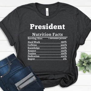 May include: A dark gray t-shirt with white text that reads "President Nutrition Facts" and lists the percentage of hard work, caffeine, knowledge, respect, passion, dedication, and regret.