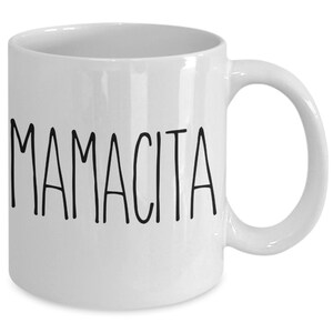 May include: White ceramic mug with the word "MAMACITA" printed in black, sans-serif font. The mug has a curved handle and a glossy finish, suitable for hot or cold beverages. A simple, yet elegant design.