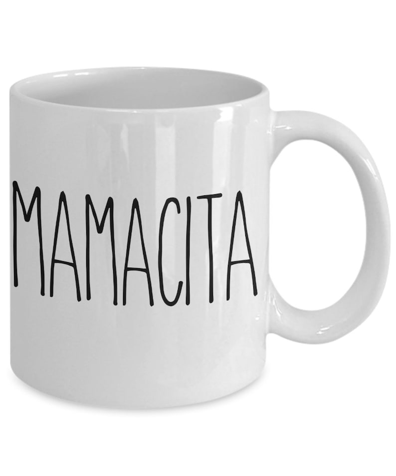 May include: White ceramic mug with the word "MAMACITA" printed in black, sans-serif font. The mug has a curved handle and a glossy finish, suitable for hot or cold beverages. A simple, yet elegant design.