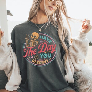 May include: A dark grey t-shirt with a colourful graphic design featuring a skeleton holding a peace sign and the text "Have the Day You Deserve."