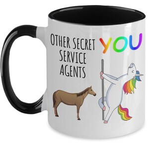 May include: White coffee mug with a black rim and handle. The mug has a graphic of a unicorn with a rainbow mane and tail, and a horse. The text on the mug reads "Other Secret Service Agents You".