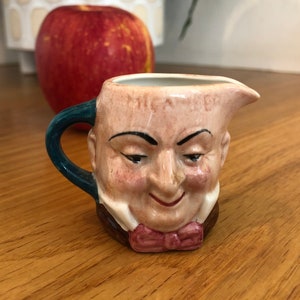 May include: A small ceramic creamer in the shape of a man's head. The creamer is painted with a realistic face and a bow tie. The creamer has a blue handle and is sitting on a wooden surface.