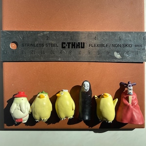 May include: A collection of small, colorful figurines, including yellow chick-like characters and a black figure, are arranged on a brown surface. A metal ruler, marked in inches and centimeters, is placed above the figurines. The figurines are approximately 1 to 2 inches in height.