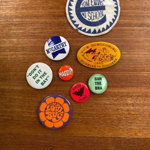 May include: A collection of vintage button pins with various designs and slogans. The pins feature a variety of colors, including red, blue, yellow, green, and orange. Some of the slogans include "Don't Do It In The Bay", "Maggie", "I'm A Love Bug", "Huelga Delano", "Ban The Bra", "Tom Evans Ski School", and "UoFW Homecoming 1963 Huskies vs Cougars".