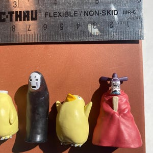 May include: A collection of small, colorful figurines, including a black figure with a white mask, a yellow chick, and a figure in a red robe. A ruler with metric measurements is placed above the figurines.