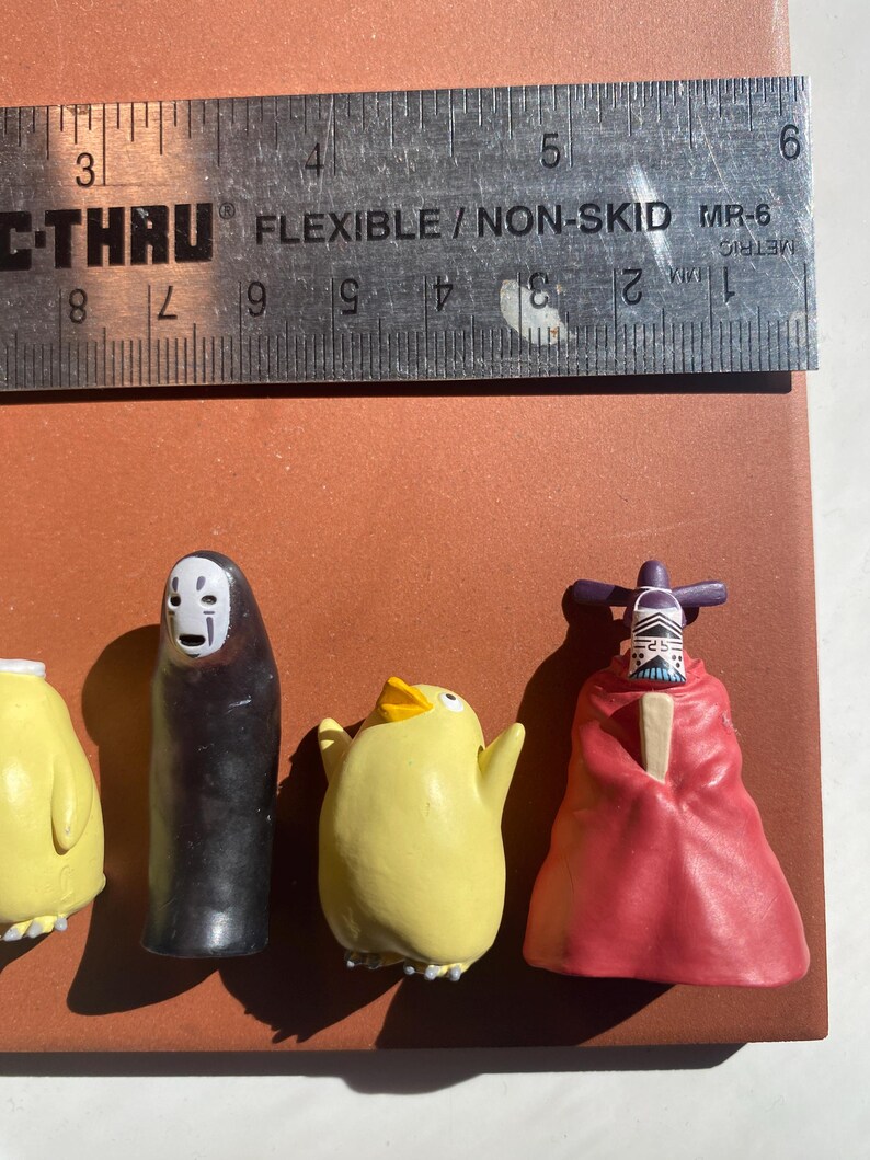 May include: A collection of small, colorful figurines, including a black figure with a white mask, a yellow chick, and a figure in a red robe. A ruler with metric measurements is placed above the figurines.