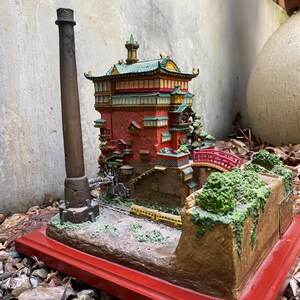 May include: A detailed miniature model of a Japanese-style building with a red base. The building has red walls, green and yellow roof accents, and a small red bridge. A tall, dark brown smokestack stands nearby.
