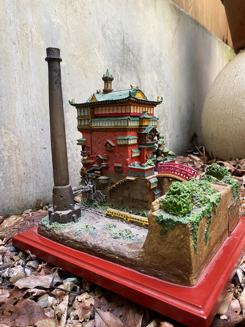 May include: A detailed miniature model of a Japanese-style building with a red base. The building has red walls, green and yellow roof accents, and a small red bridge. A tall, dark brown smokestack stands nearby.