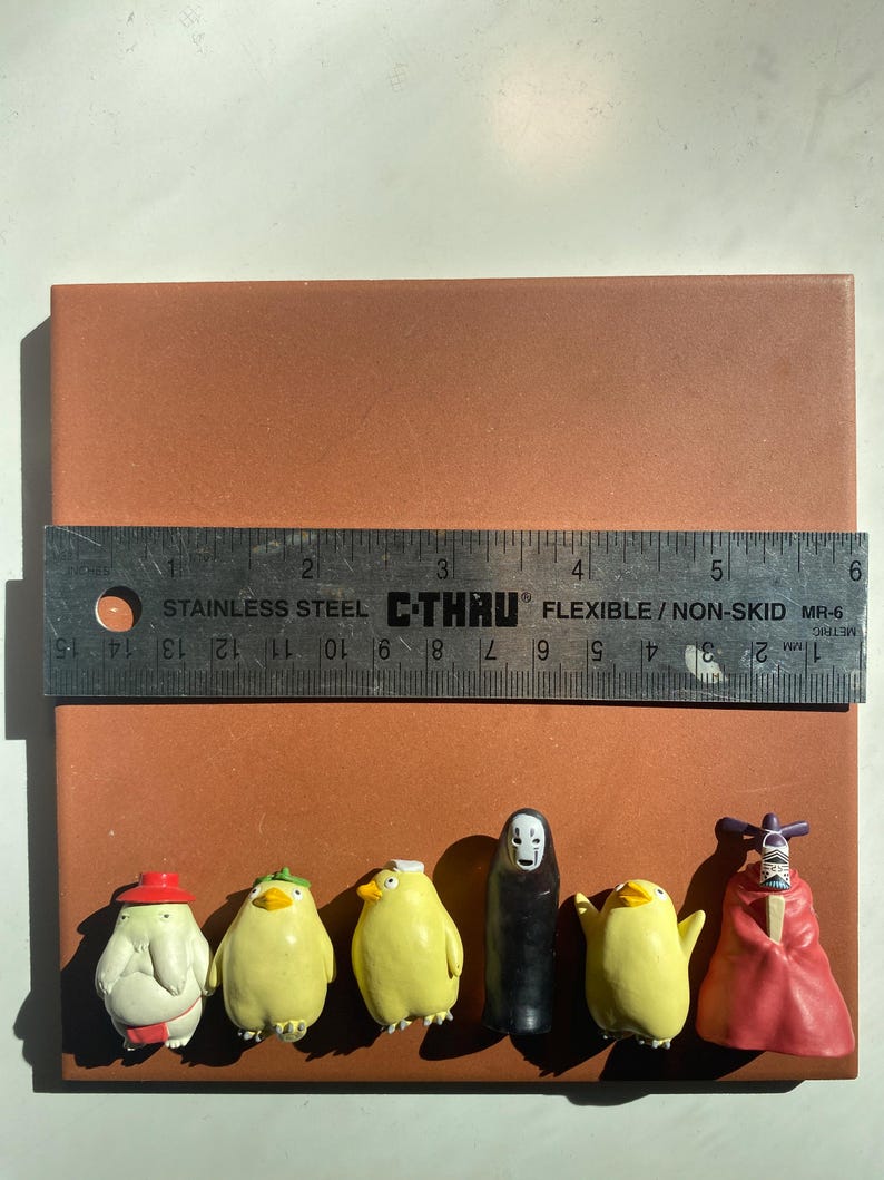 May include: A collection of small, colorful figurines, including yellow chick-like characters and a black figure, are arranged on a brown surface. A metal ruler, marked in inches and centimeters, is placed above the figurines. The figurines are approximately 1 to 2 inches in height.