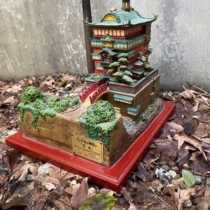 May include: A detailed miniature model of a Japanese-style building with a red bridge, green roof, and multiple levels. The model sits on a red base and is surrounded by foliage and leaves. The text on the base reads "DVD Collector's Edition".