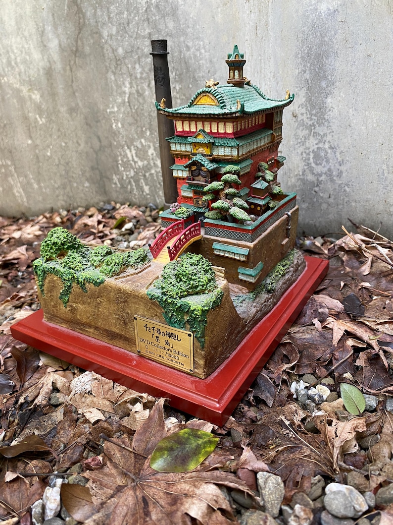 May include: A detailed miniature model of a Japanese-style building with a red bridge, green roof, and multiple levels. The model sits on a red base and is surrounded by foliage and leaves. The text on the base reads "DVD Collector's Edition".
