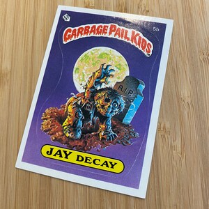 May include: A Garbage Pail Kids trading card featuring a zombie character named Jay Decay. The card has a purple background with a full moon and a tombstone that reads "R.I.P."