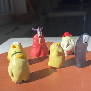 May include: A collection of miniature figurines featuring characters from a Japanese animated film. The figurines include yellow duck-like creatures, a red-robed figure, a white figure with a red hat, and a faceless figure.