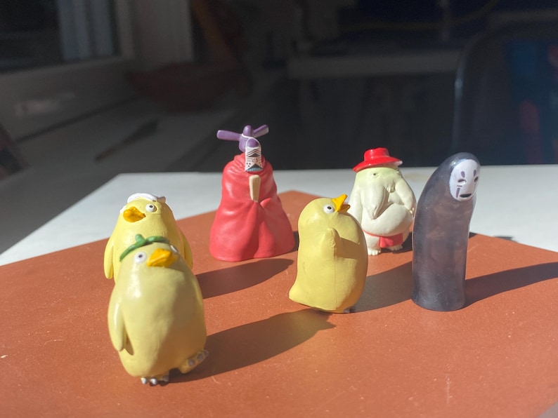 May include: A collection of miniature figurines featuring characters from a Japanese animated film. The figurines include yellow duck-like creatures, a red-robed figure, a white figure with a red hat, and a faceless figure.