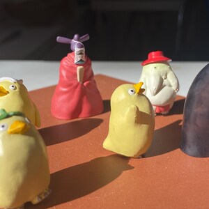 May include: A collection of colorful figurines, including yellow chick-like characters, a red-robed figure, and a tall, black, faceless character. The figurines are arranged on a brown surface, with sunlight casting shadows.