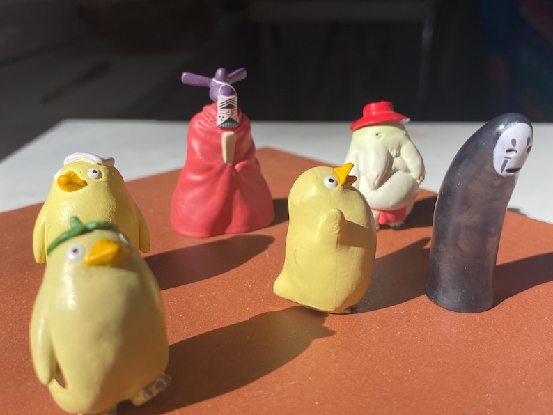 May include: A collection of colorful figurines, including yellow chick-like characters, a red-robed figure, and a tall, black, faceless character. The figurines are arranged on a brown surface, with sunlight casting shadows.