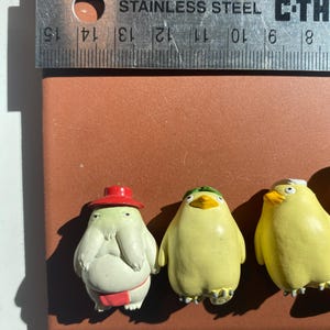 May include: Three small, colorful figurines on a brown surface. The figurines include a light green creature wearing a red hat and a red belt, and two yellow bird-like figures. A stainless steel ruler with inch measurements is above the figurines.