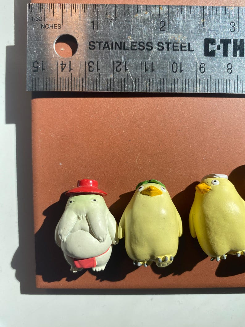 May include: Three small, colorful figurines on a brown surface. The figurines include a light green creature wearing a red hat and a red belt, and two yellow bird-like figures. A stainless steel ruler with inch measurements is above the figurines.