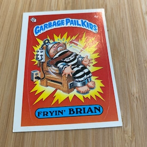 May include: A Garbage Pail Kids trading card featuring a cartoon character named "Fryin' Brian" in an electric chair. The card is red with yellow lightning bolts and the character is wearing a black and white striped prison uniform.