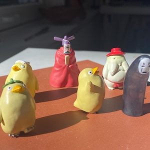 May include: A collection of miniature figurines featuring characters from a popular animated film. The figurines include yellow bird-like creatures, a red-robed figure, a white figure with a red hat, and a black figure with a white mask.
