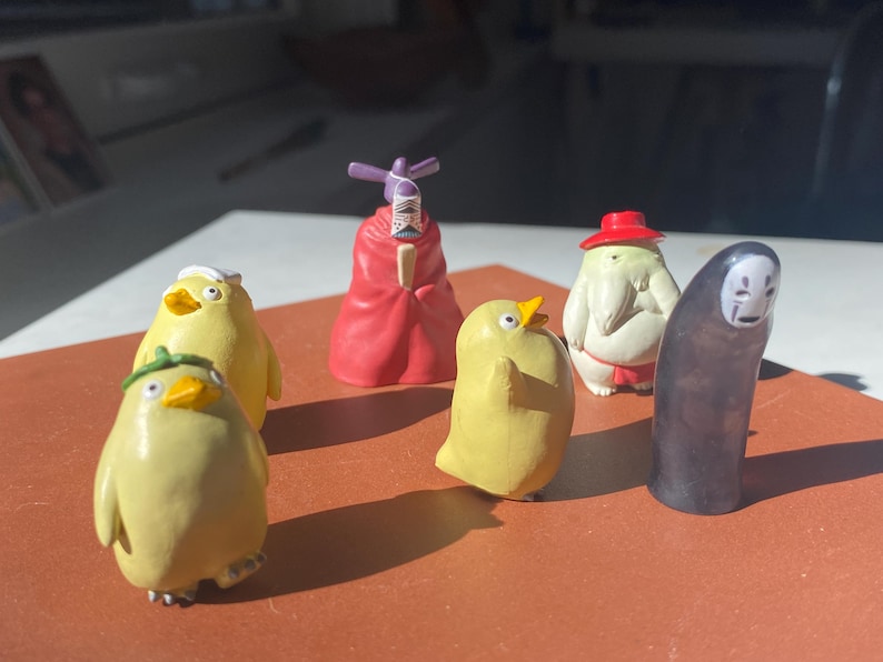 May include: A collection of miniature figurines featuring characters from a popular animated film. The figurines include yellow bird-like creatures, a red-robed figure, a white figure with a red hat, and a black figure with a white mask.