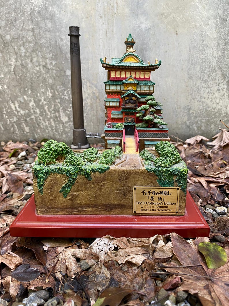 May include: A detailed miniature model of a Japanese-style building, with red, green, and yellow accents. The model includes a bridge, trees, and a small sign with Japanese text. It sits on a red base.