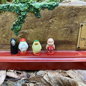 May include: A figurine set depicting characters from a Japanese animated film. The set includes a black figure, a white figure with a red hat, a yellow figure with a green hat, and a small monk figure. The base is red with a brown textured background.