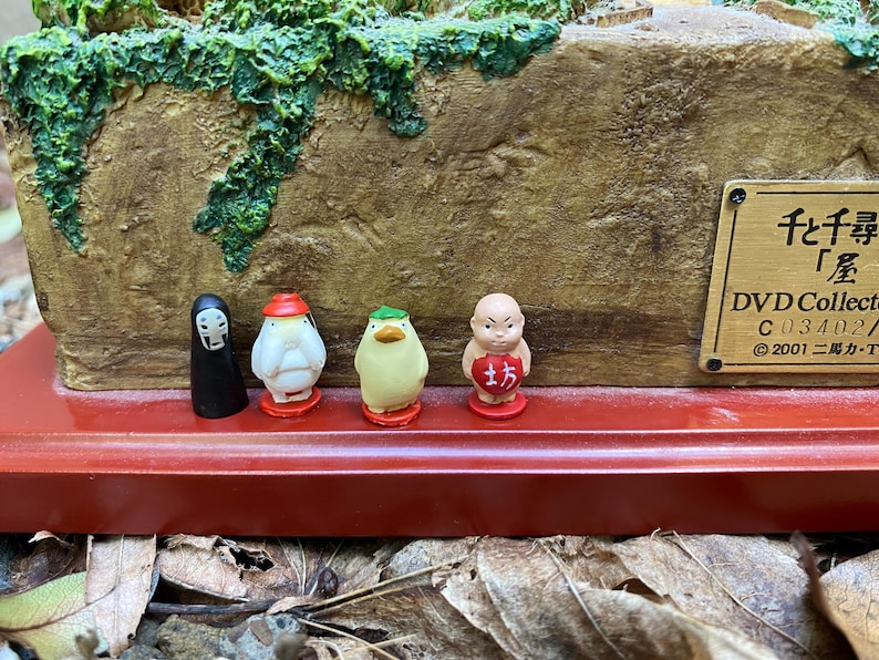 May include: A figurine set depicting characters from a Japanese animated film. The set includes a black figure, a white figure with a red hat, a yellow figure with a green hat, and a small monk figure. The base is red with a brown textured background.