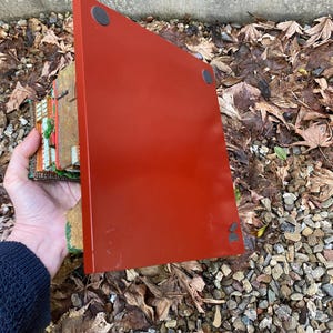 May include: A red rectangular decorative piece with black circular feet. A hand holds the piece, showing a glimpse of a detailed, colourful miniature building. The background features leaves and small stones.