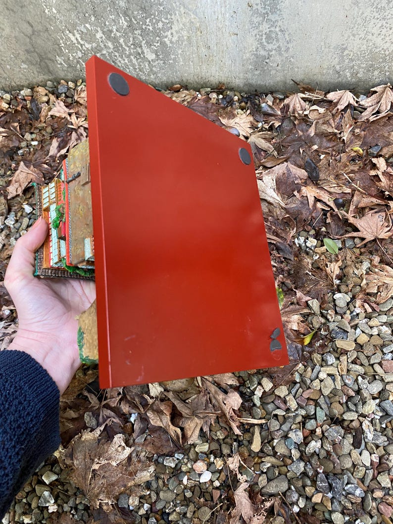 May include: A red rectangular decorative piece with black circular feet. A hand holds the piece, showing a glimpse of a detailed, colourful miniature building. The background features leaves and small stones.