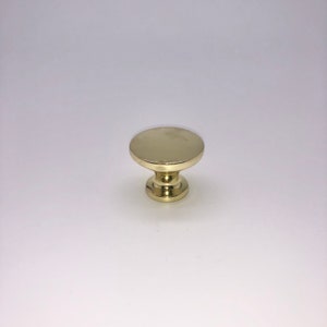 Pode incluir: Um bot&atilde;o de arm&aacute;rio redondo, dourado, com uma parte superior plana e uma base ligeiramente elevada.