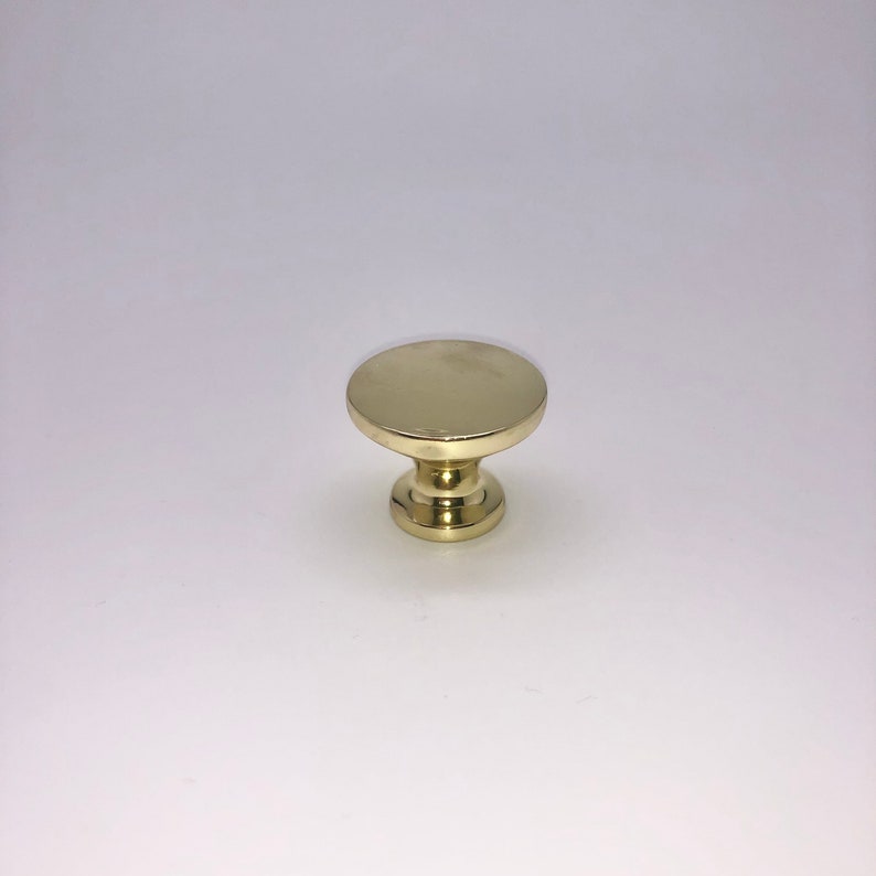 Pode incluir: Um bot&atilde;o de arm&aacute;rio redondo, dourado, com uma parte superior plana e uma base ligeiramente elevada.