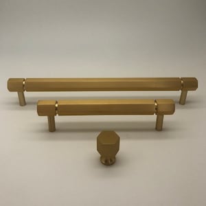 Op de afbeelding: Drie gouden kastbeslagknoppen. Twee zijn rechthoekig met zeshoekige uiteinden en één is een ronde knop met een zeshoekige vorm.