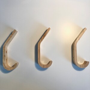 May include: Three wooden wall hooks in the shape of a 'J' with a rounded top. The hooks are made of light brown wood and are mounted on a white wall.