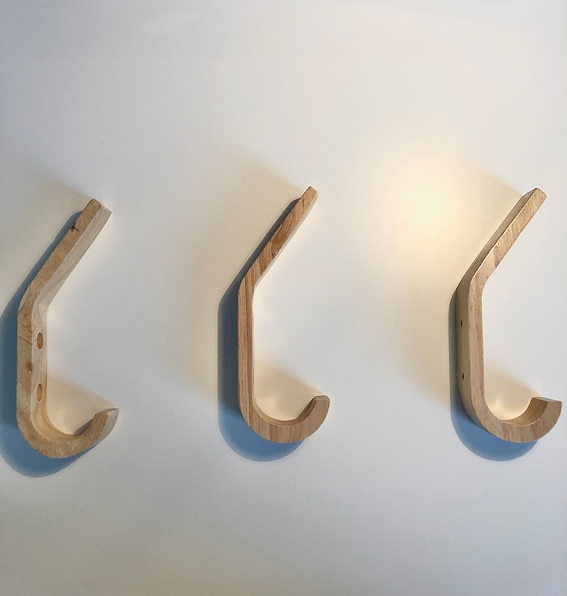 May include: Three wooden wall hooks in the shape of a 'J' with a rounded top. The hooks are made of light brown wood and are mounted on a white wall.