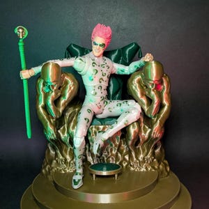Könnte beinhalten: Figur des DC Comics Charakters Riddler, sitzend auf einem goldenen Thron. Die Figur ist hellgrün mit Fragezeichen, rosa Haaren und einem grünen Stab. Der Thron hat zwei goldene Figuren in Denkpose.
