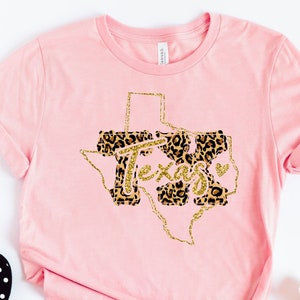 May include: Pink t-shirt with a gold glitter outline of the state of Texas. The inside of the state is filled with a leopard print pattern and the word "Texas" is written in gold glitter.