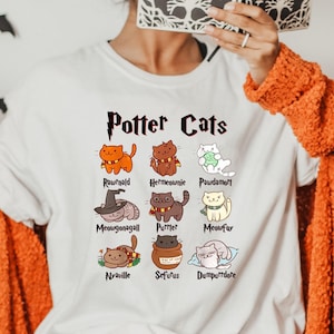 May include: White t-shirt with a graphic of cats representing characters from the Harry Potter series. The text "Potter Cats" is at the top of the graphic. The names of the characters are listed below each cat.