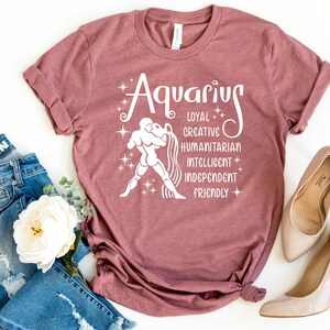 May include: A mauve short-sleeved t-shirt featuring white screen-printed text that reads Aquarius, Loyal, Creative, Humanitarian, Intelligent, Independent, Friendly, alongside a graphic of a figure pouring water and several small star icons.