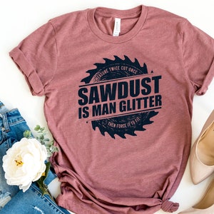 Pu&ograve; includere: Una maglietta color marrone rossiccio con una grafica nera di una lama da sega che recita "Measure Twice Cut Once Sawdust Is Man Glitter Then Force It To Fit".