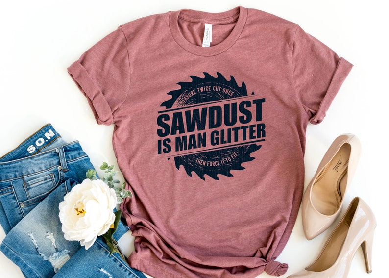 Pu&ograve; includere: Una maglietta color marrone rossiccio con una grafica nera di una lama da sega che recita "Measure Twice Cut Once Sawdust Is Man Glitter Then Force It To Fit".