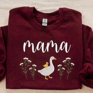 May include: A maroon sweatshirt with the word "mama" in white script. Below the text is an embroidered design of a goose with a yellow duckling on its back, surrounded by white flowers with green stems.