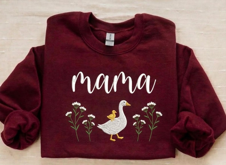 May include: A maroon sweatshirt with the word "mama" in white script. Below the text is an embroidered design of a goose with a yellow duckling on its back, surrounded by white flowers with green stems.
