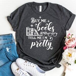 Puede incluir: Una camiseta gris oscuro con el texto "Buy me books and tell me I'm pretty" en blanco. El texto est&aacute; rodeado por una pila de libros y un par de gafas.