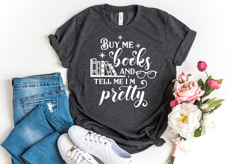 Puede incluir: Una camiseta gris oscuro con el texto "Buy me books and tell me I'm pretty" en blanco. El texto est&aacute; rodeado por una pila de libros y un par de gafas.